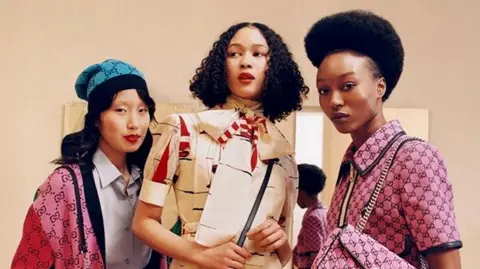 Bolade Banjo A Gucci advert staring three people all wearing the designer. Lisa is on the right hand side wearing a pink suit and matching handbag and has an afro. The middle woman has dark brown shoulder length curly hair and is wear a beige outfit with a neckerchief. The third woman is wearing a bright pink cardigan and blue hat.