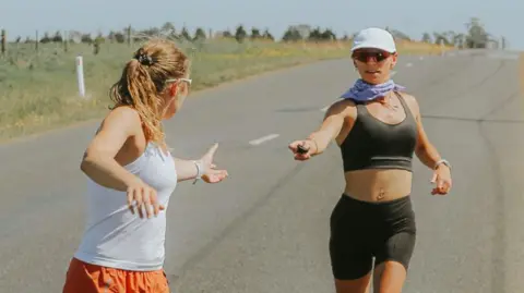 Megan Chapple hands a Garmin device to another team member completing the run in Tasmania. The terrain is flat and it is a sunny day. A car is visible to the right.