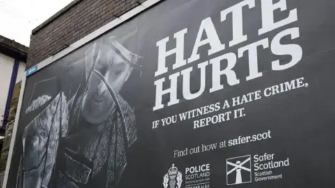 A black and white outdoor advertising poster from 2024 for a Scottish government campaign to encourage people to report hate crimes 