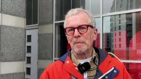 Sofía Luis-Hobbs/BBC A man with red circular framed glasses looks at the camera. He wears a white T-shirt under an unbuttoned brown and black striped shirt which sits beneath a red and navy zip up raincoat.