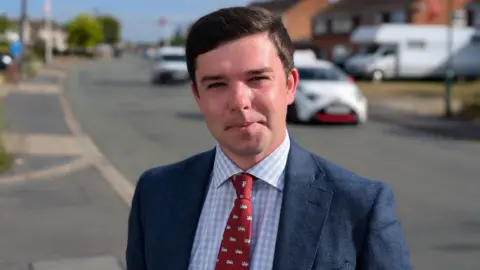 BBC Alex Wagner has brown hair and is standing by a road. He is wearing a navy suit jacket, a blue and white check shirt and a red tie with white lions on it.