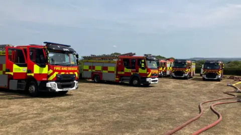 Six fire engines on the grass which appears yellow and dry
