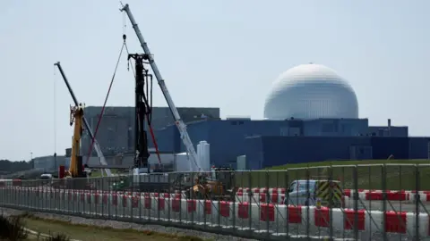 The large white dome of the Sizewell B nuclear power plant is the backdrop of the picture which has construction work and cranes in the foreground. The white dome sits on large square grey coloured buildings. 