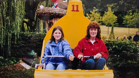 Collect/PA Real Life An older photo of Smith and her mum when Smith was a teenager. They are sitting on a large yellow seat that looks to be part of a ride. They have a metal bar across their laps. Smith has shoulder-length brown hair and is wearing a blue coat. Next to her is her mum who has curly red-brown hair and is wearing a red jacket