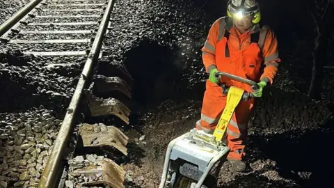 A rail engineer works at night on repairs to a line in Devon. He is wearing orange high-vis overalls and has a light on his helmet. He also has a piece of equipment in his hands.