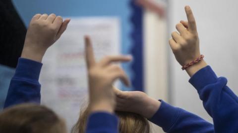 Children with their hands up wearing blue jumpers in a classroom