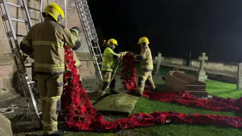 Twisted Stitches Three firefighters wearing full kit - beige coats and trousers with yellow reflective material, and yellow helmets. They're holding the ends of the poppy cascade and are about to climb two metal ladders propped up against the church bell tower. It's pitch black outside, so it taken late at night, but flood lights have been set up. 