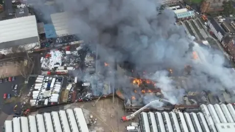 West Midlands Fire Service The large fire is captured from above. Large plumes of smoke and flames can be seen coming from a building. The smoke fills the picture.