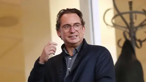 Simon Sadler is wearing a dark jacket and shirt and is pictured inside a room with a hat stand in the background.