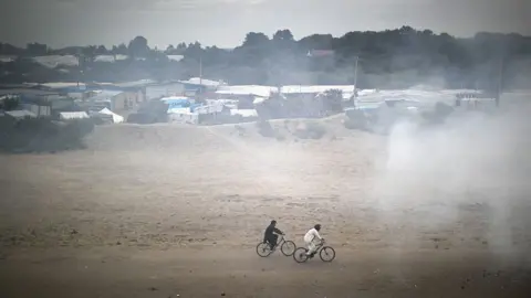 Getty Images Migrants cycle through smoke from a fire burning rubbish at the Jungle migrant camp on September 6, 2016 in Calais, France.