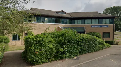 The outside of Elizabeth House, at Fulbourn Hospital. It is an angular brick building, with a line of windows on the top floor. In the foreground is a big green bush and in the distance, on the right, is a police car parked up.