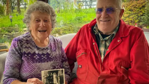 Marilyn and Pete in their lounge, with Marilyn holding a wedding picture