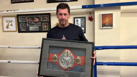 Stephen Brown/BBC A man in a black t-shirt is holding a kickboxing title belt, encased inside a frame. He is standing inside a boxing ring.