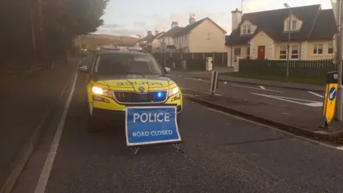 BBC Police car behind sign reading "POLICE ROAD CLOSED"