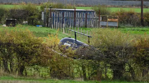 SWNS A black helicopter on its side in a field, partly concealed behind some bushes.