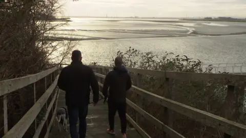 Silhouettes of two men and a dog walking down a wooden plank footpath towards the Mersey estuary