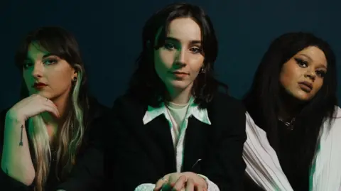 Sarah Ward CHERYM , made up of L-R Alannagh Doherty, Emer McLaughlin and Hannah Richardson. The band are dressed in monochrome outfits and sit posing on a black sofa against a dark blue backdrop.