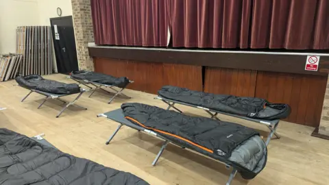 West Northamptonshire Council Camp beds with a sleeping bags on them in the hall. There is a large red curtain in the background