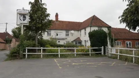 A white building with a sign on the left hand side saying "The White Hart". There's vegetation and an empty car park in front of it.