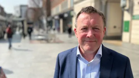Devon County Councillor Neil Stevens in a blue and white fine-striped shirt and a blue suit jacket in Exeter High Street