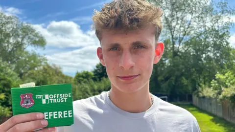 A close up image of Luca Sinclair who has blond hair and blue eyes. He is in his garden holding his green referee arm band, which reads "The Offside Trust, Referee". He is wearing a grey T-shirt with a silver chain around his neck. 