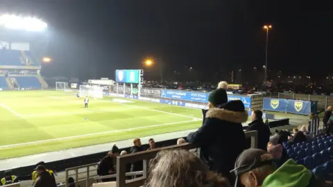 Malcolm Townrow Kassam Stadium from the stands. The pitch is green and there are players on it.