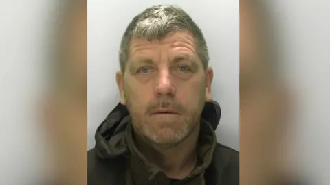 A policy custody image of Scott Herbert. He is middle aged with short grey hair and grey stubble. He wears a dark green jumper and black anorak. 