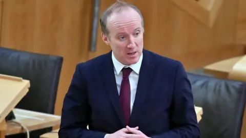 Getty Images Michael Marra in a dark blue suit and burgundy tie stands and speaks in a chamber with wooden desks and black chairs visible around them.