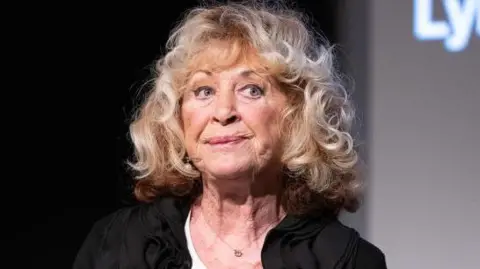 La Plante pictured at an event in 2021, looking away from the camera with a pensive expression on her face, wearing a black top and big blonde curls.