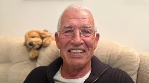 Tim Shepherd grins at the camera while sitting on a sofa. He has short, white hair and wears square, thin-framed glasses, a white T-shirt and a dark, knitted, zipped top.