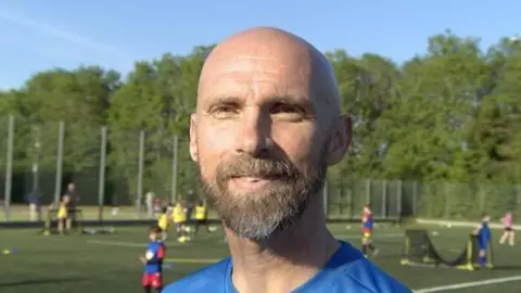 Steve Hubbard/BBC Close-up image of Luke Chadwick. He has a bald head and beard and is smiling at the camera. He is wearing a blue T-shirt. Behind him are some pitches where children are playing football.