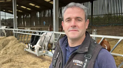 Ben Yates is standing in his barn with several cows eating behind him. He is wearing a black gilet and navy shirt. He has short grey-brown hair.