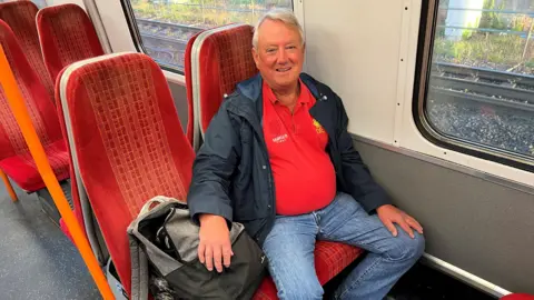 A man sitting on a train, smiling for the camera. There is a rucksack next to him.
