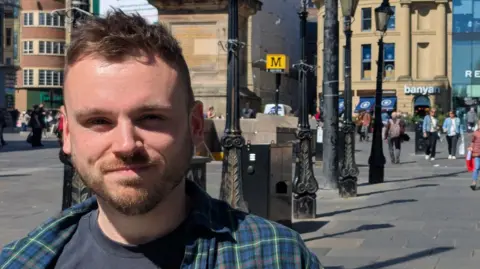 Kieran Carter, a man in his late 20s with short, brown hair and a stubble beard. He is wearing a blue tartan shirt. He is stood in Newcastle's city centre near a metro station.