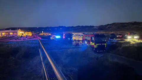 BBC A fire engine and other emergency vehicles parked. To the left are caravans with people stood around. There are large hills in the background. The sky is a dark grey. 