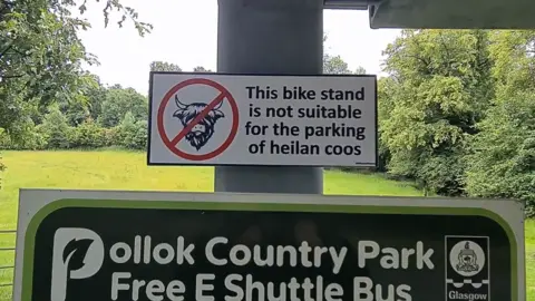 @ASignOfHumour A photo from Pollok Park where Highland Cows are a popular attraction. The sign has the head of a highland cow in a circle and a line through it to show they are prohibited. The text reads: This bike stand is not suitable for parking heilan cows.