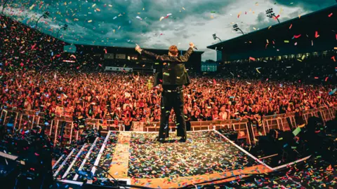 IantheWarlow A man stands on a stage in front of thousand of people holding his arms aloft while clutching a microphone as confetti and ticker tape falls all around him.