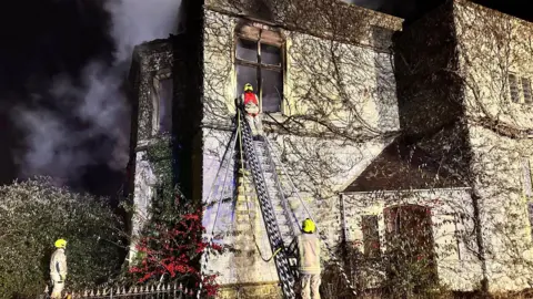 OFRS Firefighters tackling the Bignell House fire during the night. Smoke is seen coming from the roof. One firefighter is seen on a ladder next to the building, while two other crew members are supporting them from below with a fire hose.