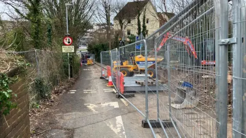 A road blocked off with temporary metal fencing. There is a digger to the right. There is a give way sign on a lamppost. In the background are trees and houses. 