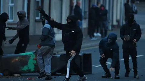 Getty Images Six men wear black coats and black tracksuit bottoms. They all wear black balaclavas as they throw stones, bricks and other objects. A group of people, out of focus, can be viewed looking on in the background. Two black wheelie bins lie beside the individuals throwing the rocks.
