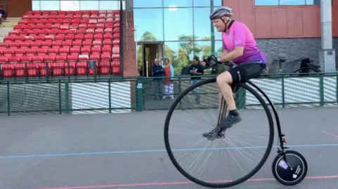 Rolf cycles past on a penny farthing on a cycle race track.