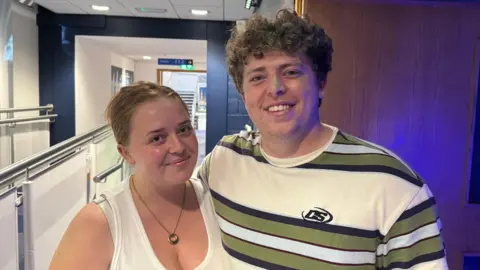 Abbie and Scott pose for pictures in the hallway of the Armagh Planetarium. One is a young woman with her hair tied back, she is smiling at the camera and is wearing a white tank top and a silver, circle necklace. Scott, on the right, has curly hair and is smiling at the camera. He is wearing a white shirt with green, blue and black stripes.