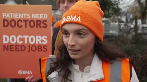 Dr Heather Gunn has long, curly brown hair under her orange BMA beanie hat. She is wearing an orange fluorescent jacket and standing on the picket line, next to a sign that reads "Patients need doctors. Doctors need jobs."