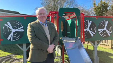 Joanna Taylor/BBC Councillor Peter McDonald stands in front of the climbing frame. The green climbing frame has 'wings' bordered in red with propellers painted on. A slide comes out from the front of the page, after a wooden crawl space. Cllr McDonald wears a green blazer, beige jumper, blue checked shirt and yellow and blue striped tie. He is smiling and stands with his hands clasped in front of him. 