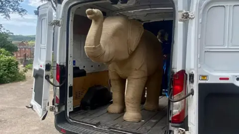 The large model of Ropey, a model elephant, is standing in the back of a van, which is open with its back doors open.
