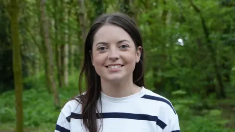Kathryn is stood in a wooded area, she is wearing a white striped jumper and has long, dark hair.