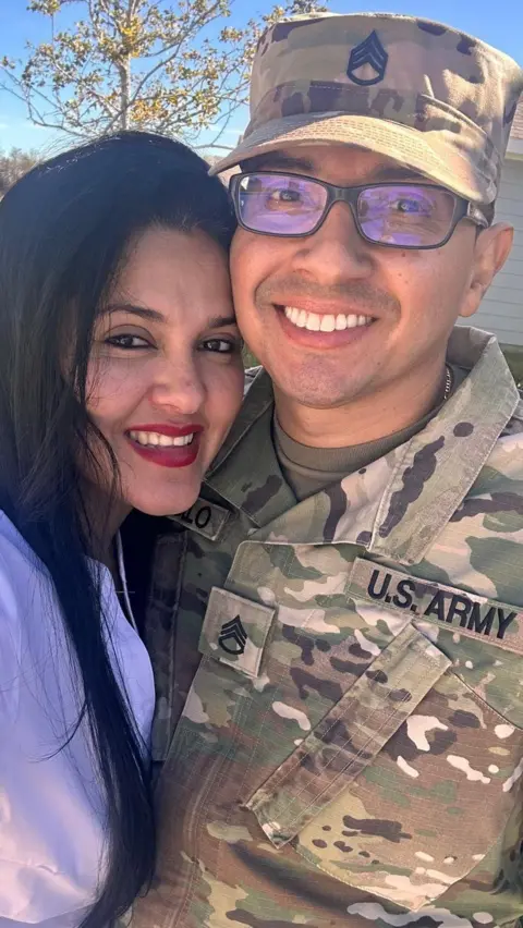 Courtesy of Wilmer Trujillo Retired Staff Sergeant Wilmer Trujillo and his wife, Arelys Martinez, in an undated photo. It is a selfie photo; Arelys smiles and wears a white blouse. Sgt Trujillo is wearing his Army uniform, looking at the camera and smiling.
