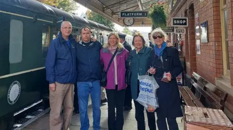 Five people, wearing coats and trousers - three women and two men - stand beside a train on a train platform