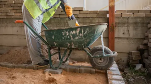 The lower half of a man in grey jogging bottoms and a yellow high visibility jacket standing beside a green wheelbarrow, resting on some grey paving stones sitting in sand.