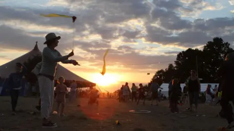 Towersey Festival A man wearing a hat waves two sticks in the air. The sticks have yellow ribbons on them. In the background, the sun is setting and there is a large festival tent. A number of punters can be seen walking around, although most of them are silhouettes.